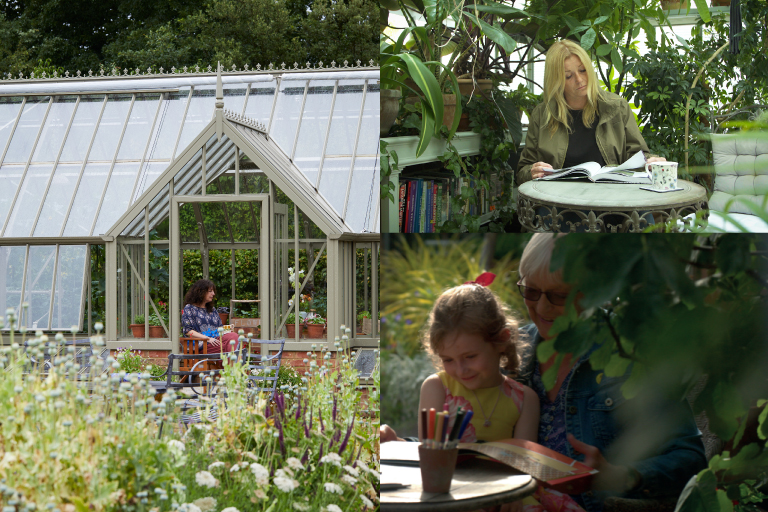 A woman reading a book at a small table inside a plant-filled greenhouse, with a grandmother and child drawing together